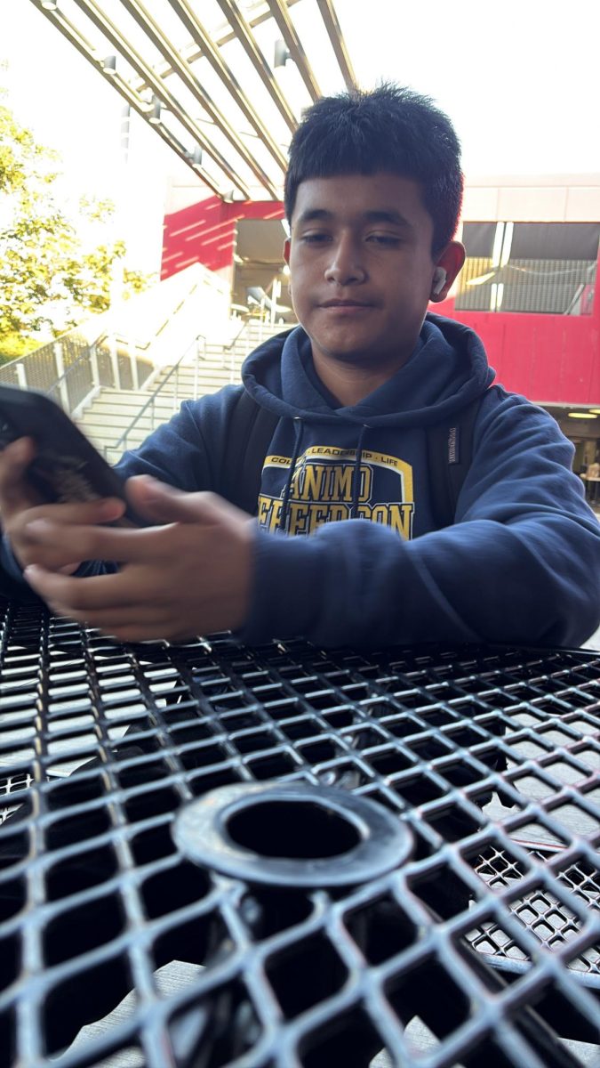 Jose sits down on the bench on the JMS patio playing on his phone. 